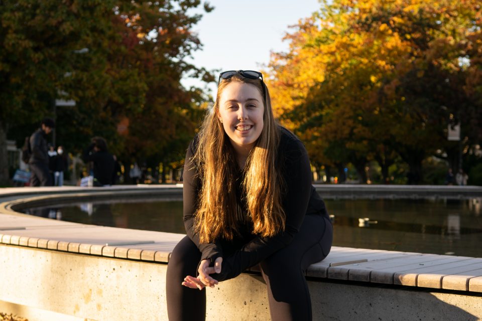 Photo of Meghan Cooke sitting outside, near a fountain, on the UBC campus.
